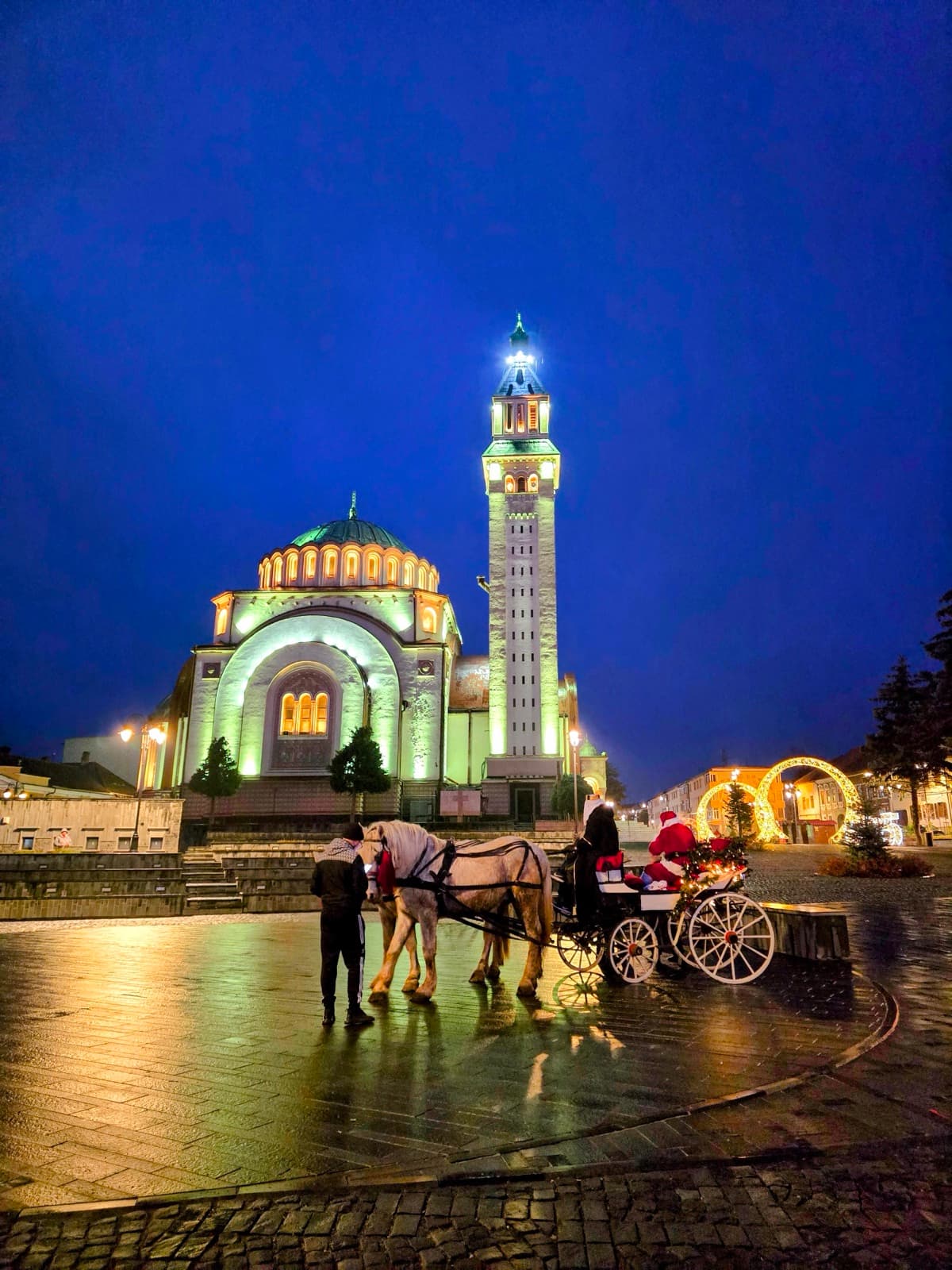 Orăștie's Orthodox cathedral lit up at dusk, with a horse-drawn carriage in the festive square
