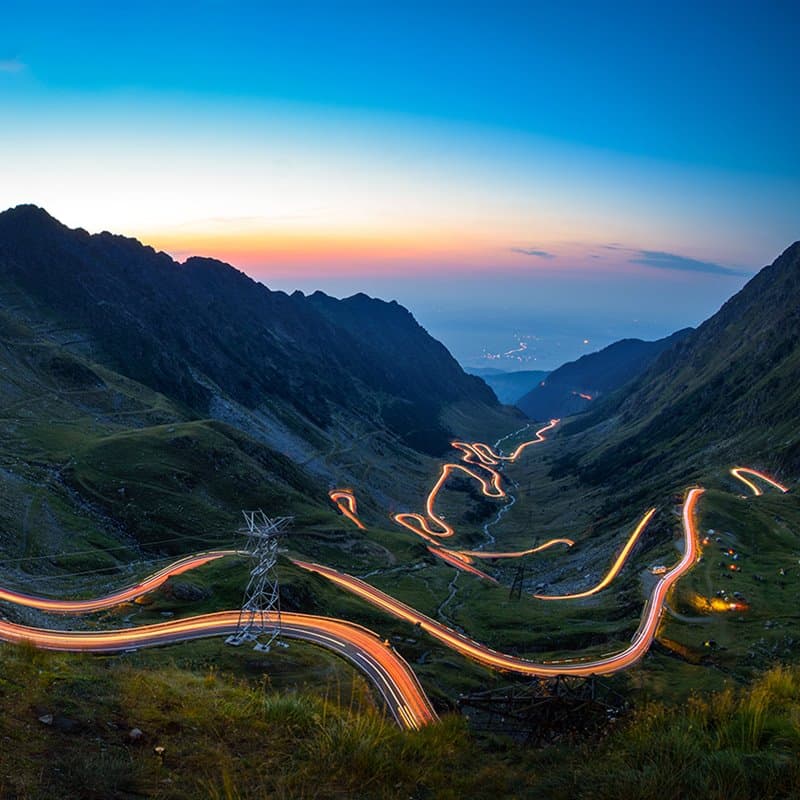 The Transfăgărășan's hairpin bends glowing with car light trails at dusk, high in the Carpathians
